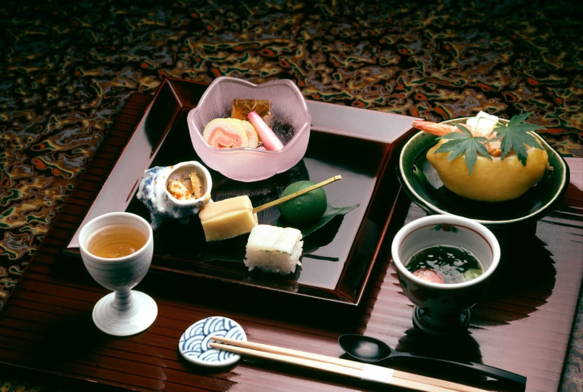 伝統的な懐石料理のセットが赤いお膳に並ぶ。焼き魚、ご飯、お椀が配置され、茶の湯の簡素で均衡のとれた美学を表現している。